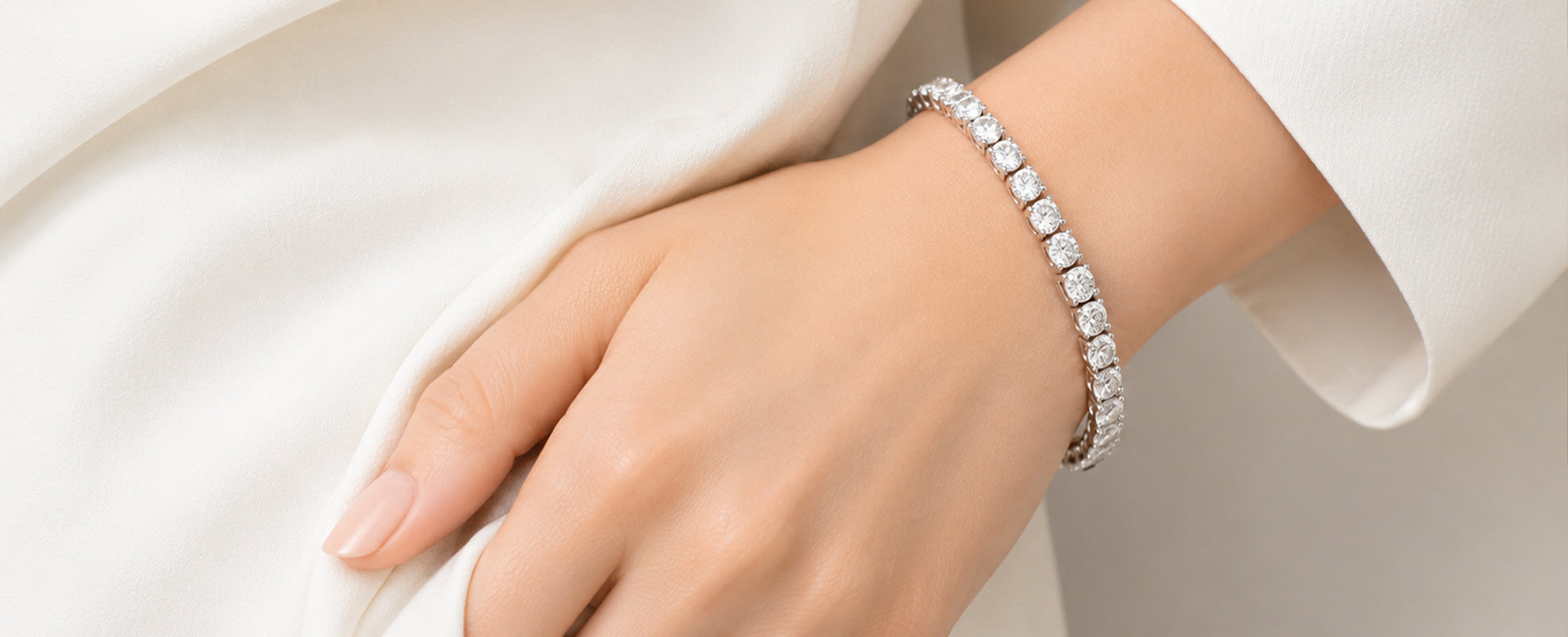 A close-up of a person's hand and wrist wearing a silver tennis bracelet with clear gemstones, styled with a white sleeve.