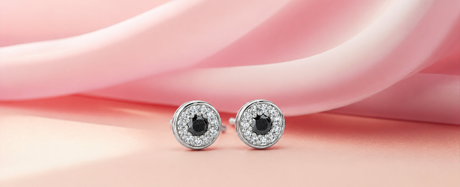 A pair of round stud earrings with silver-toned settings, surrounded by small clear stones and featuring a central black stone, displayed on a soft pink background.