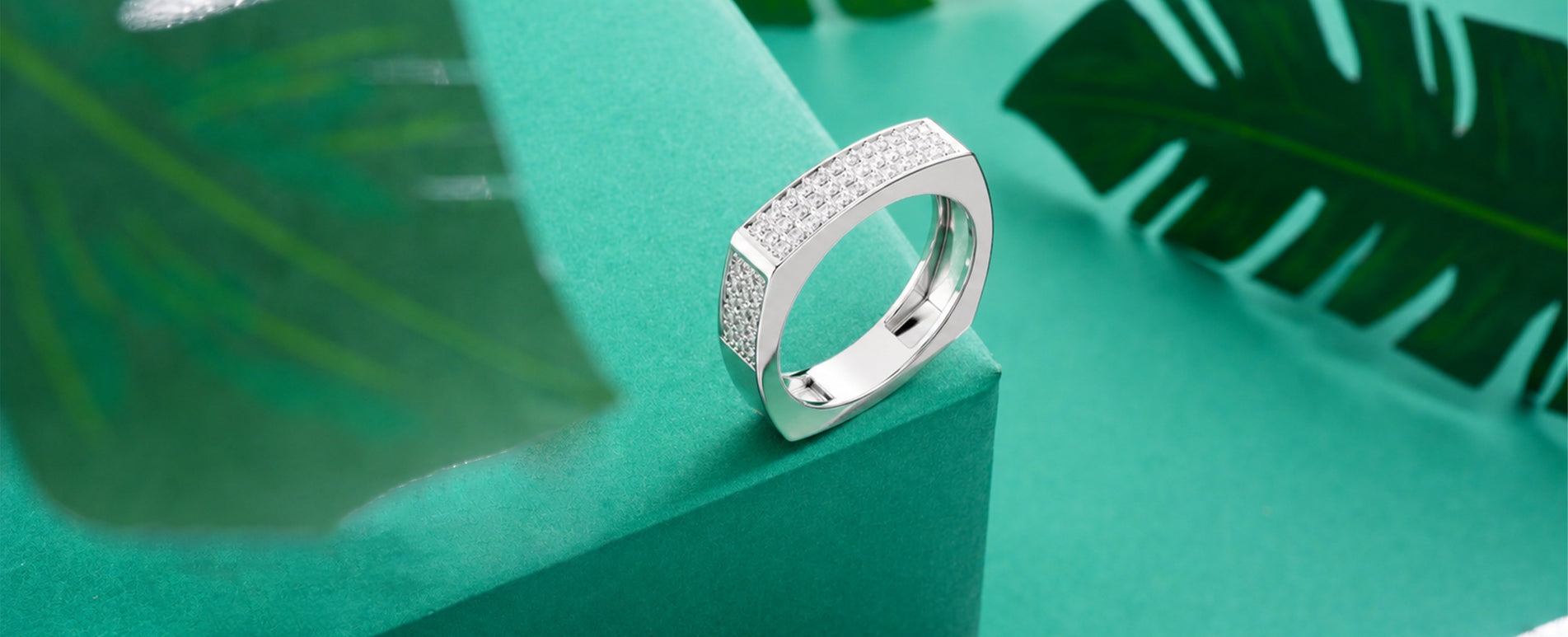 A silver band ring with pavé-set clear stones, standing upright on a teal-green block with tropical leaves in the background.